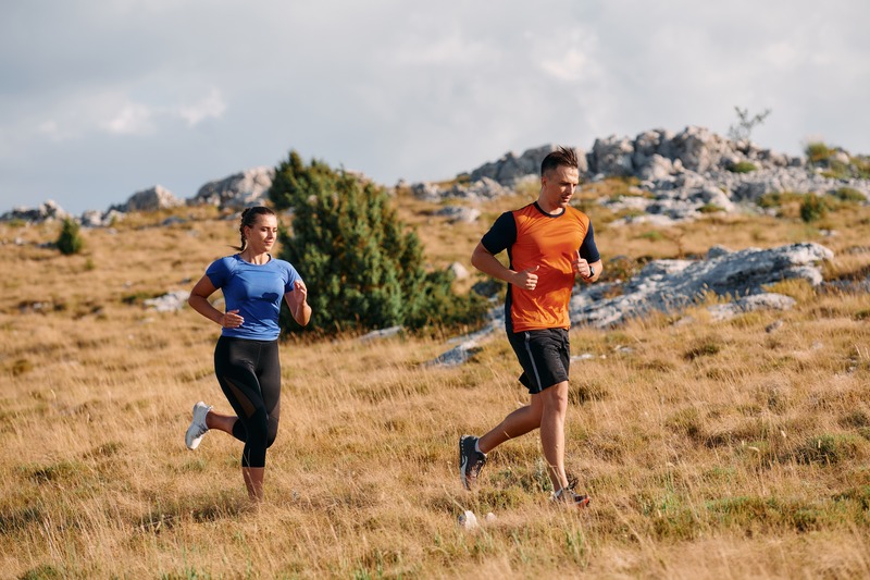 A man and a woman running on a grassy trail in a rocky landscape.